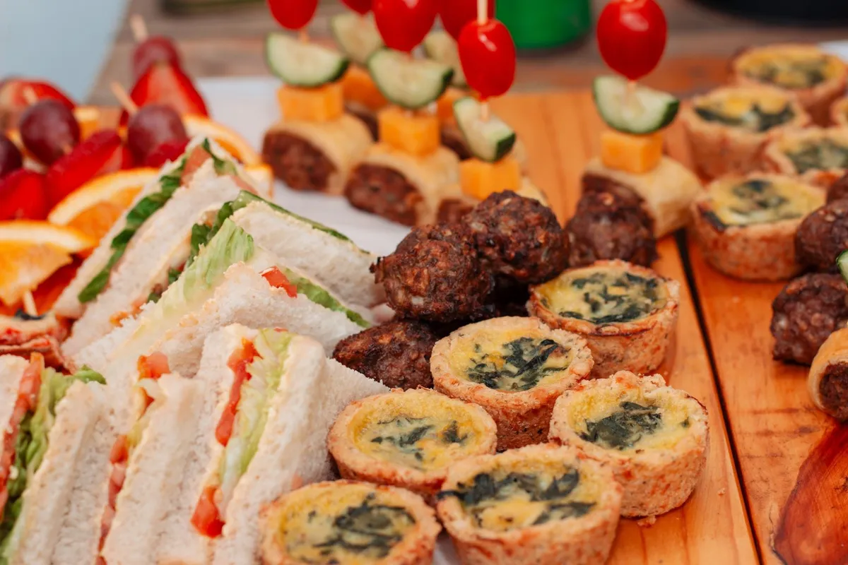 A wooden platter filled with assorted finger foods like sandwiches, quiches, and kebabs for casual dining.